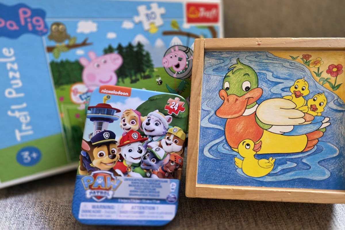 Kolorowe puzzle dla dzieci: Peppa Pig, Psi Patrol i drewniana układanka z kaczką i kaczątkami na tle sofy.