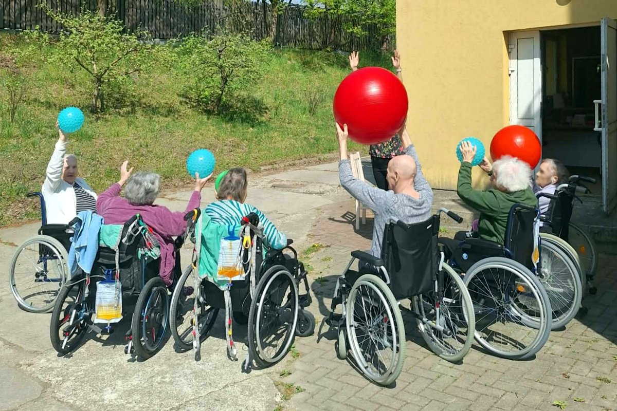 Seniorzy na w&oacute;zkach inwalidzkich ćwiczą na świeżym powietrzu z kolorowymi piłkami przed budynkiem.